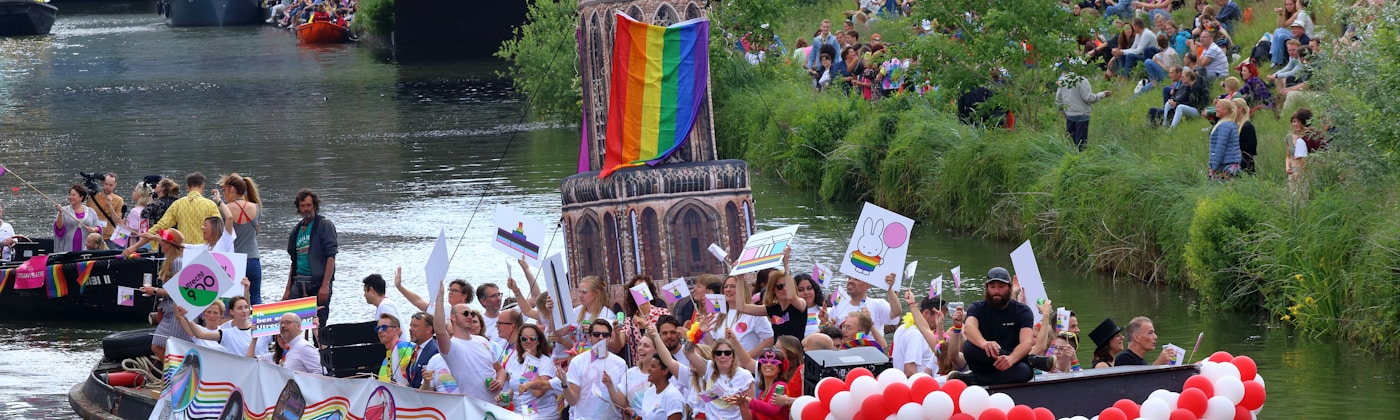 A large crowd of LGBTQ+ community members celebrating Pride on a float in a canal, waving rainbow flags — the community WanderSafeMesh is built to protect